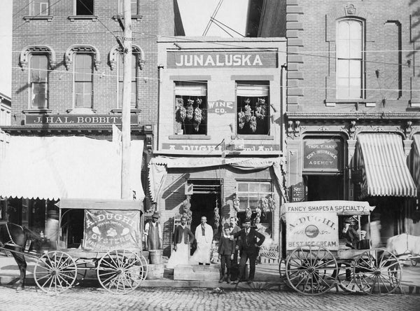 Dughi Store at 235 Fayetteville Street in Raleigh, 1895. In front of the store is an oyster wagon pulled by horse Nancy and an ice cream wagon pulled by horse Jno. From left: L. C. Bell, Nathan Sledge, Tounie Dughi, Antonio Leo Dughi, John J. A. Dughi. Adjacent businesses include J. Hal Bobbitt and the Thos. T. Hay General Insurance Agency. Courtesy State Archives of North Carolina / #PhC.166.4