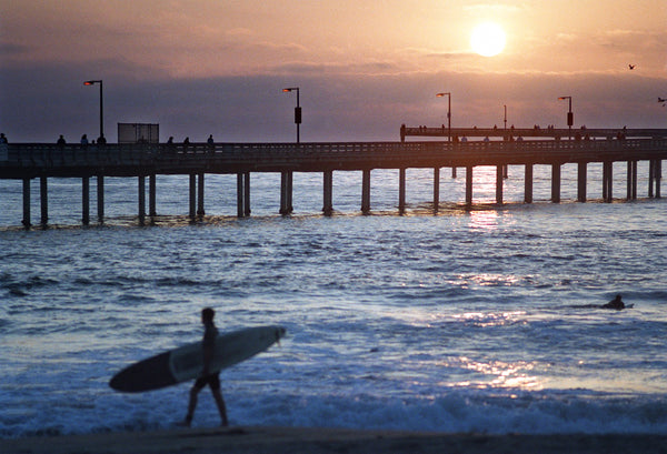 A surfer walks along the sand in Ocean Beach with the pier and the setting sun in the background, June 20, 1996. John Gastaldo / The San Diego Union-Tribune