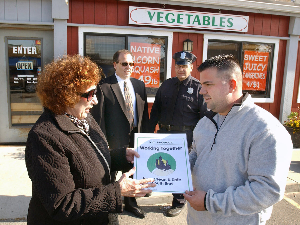 Coordinator Angelina R. Florian, left, hands a sign to Antonio Calabrese, owner of AC Produce at 489 Main St., Springfield, Tuesday. Looking on at rear are South End Citizens Council Inc. President Leo A. Florian, left, and Community Policeman Robert Jacobson. Courtesy Mark M.Murray / The Republican