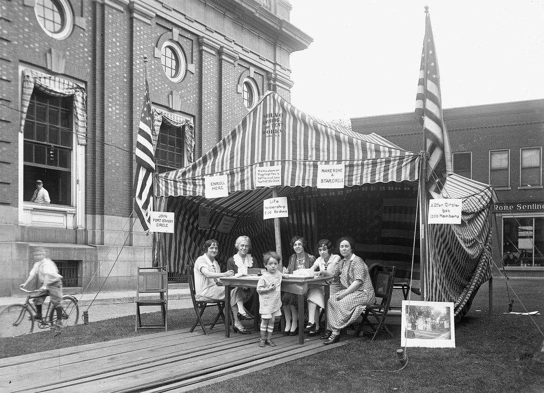 Five women sitting behind a table and one young boy in a tent between the Post Office building and the Rome Sentinel building in the background, circa 1925. A large photo of the Rome Club on East Dominick Street is leaning on the right side of the tent and a Post Office employee is in the window on the left. Courtesy Rome Historical Society