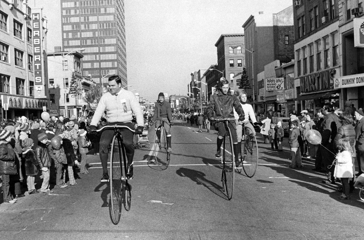 Downtown Merchants Parade, November 29, 1975, looking north on Elm Street. Herbert’s Music Store is seen on the left side of the image (west side), followed by Stark Street, and in the background Hampshire Plaza. A crowd of people gather on both sides of the street watching the parade as four men on high wheel bicycles ride by, John Dubuclet is on the left rear and Frank Skrzyzowski right rear. Courtesy Manchester Historic Association