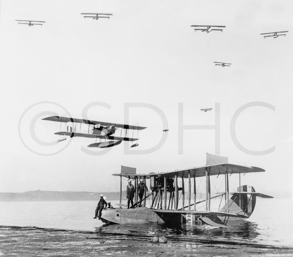 Men stand on a Navy seaplane in San Diego Bay off North Island Naval Air Station with JN-9 airplanes flying above. San Diego’s mild weather made it an ideal test ground for naval aviation, beginning in 1911. San Diego History Center (#6091)