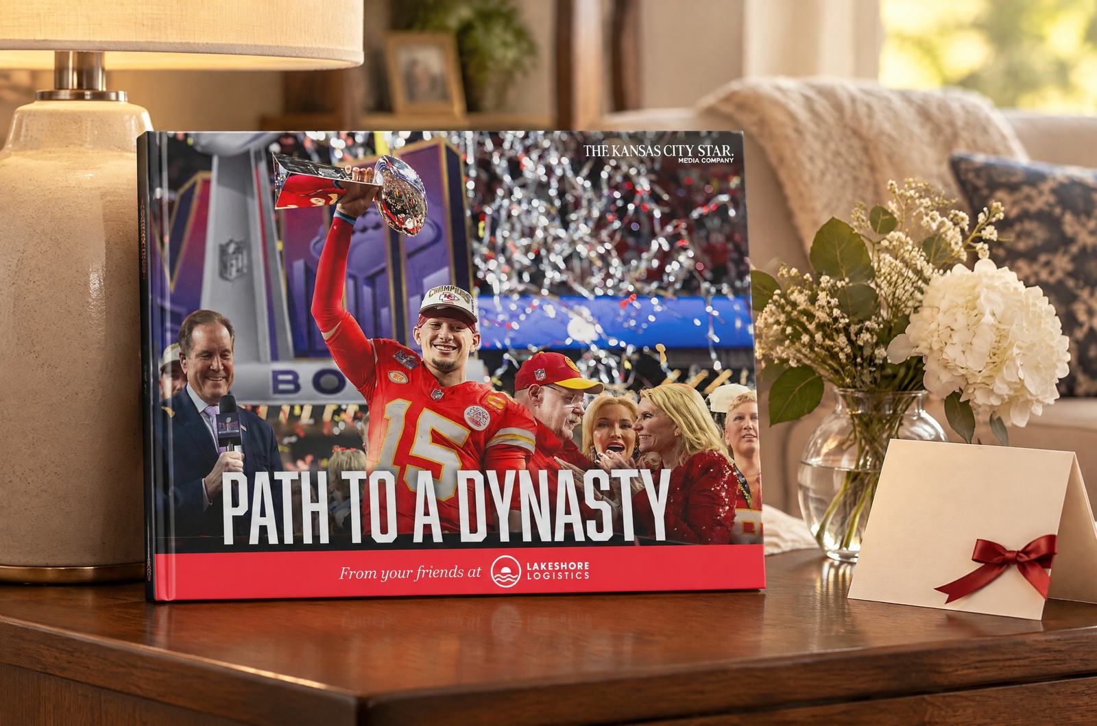 The book “Path to a Dynasty”, with the Lakeshore Logistics logo on red along the bottom of its cover, standing upright on a wooden side table beside a greeting card and flowers in warm evening light.
