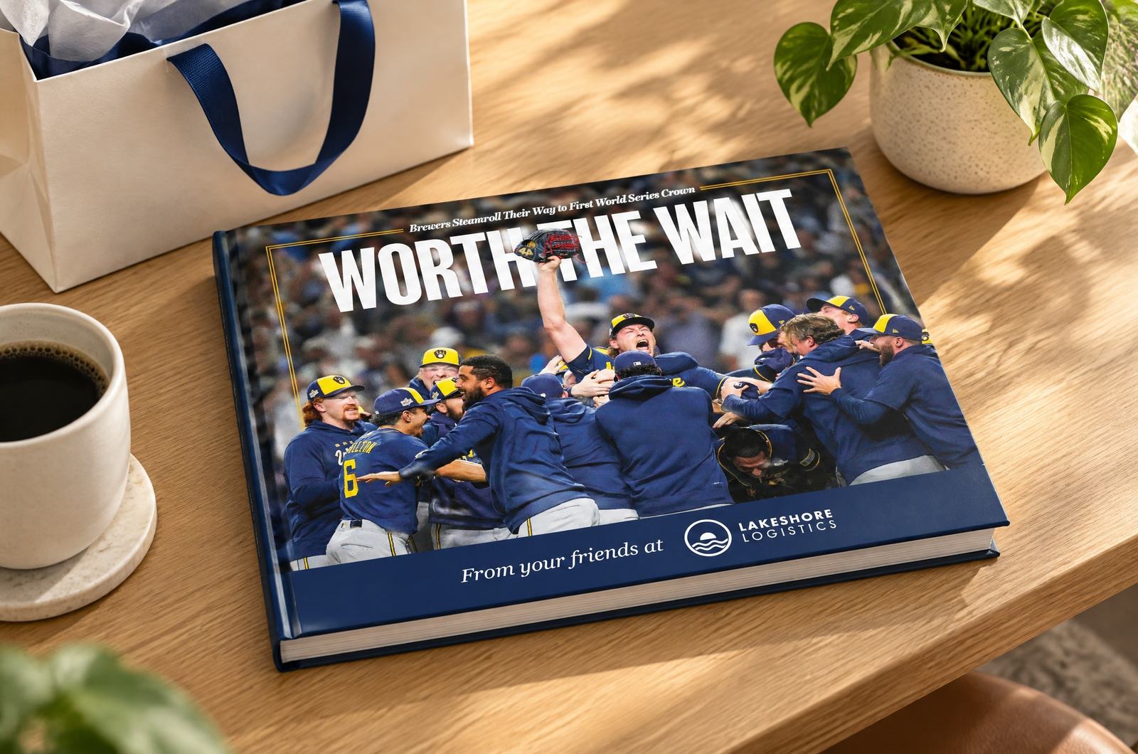 A landscape hardcover with “From your friends at” and the Lakeshore Logistics logo on navy along the bottom of its cover, lying flat at a mild angle on a light-oak desk beside a cream gift bag with a navy ribbon, a coffee cup and a small plant.